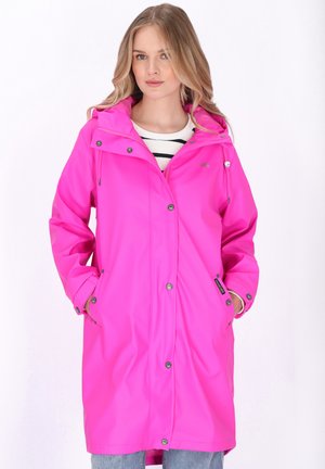 Femeie purtând o pelerină de ploaie roz aprins cu glugă, cu mâinile în buzunare, peste o cămașă în dungi și blugi albaștri, stând în picioare pe un fundal alb.