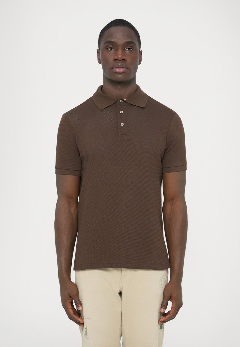 Hnedé polo tričko s krátkymi rukávmi, zapínaním na gombíky a textúrovanou látkou. Kombinované s nohavicami svetlej farby a jednoduchým pozadím.