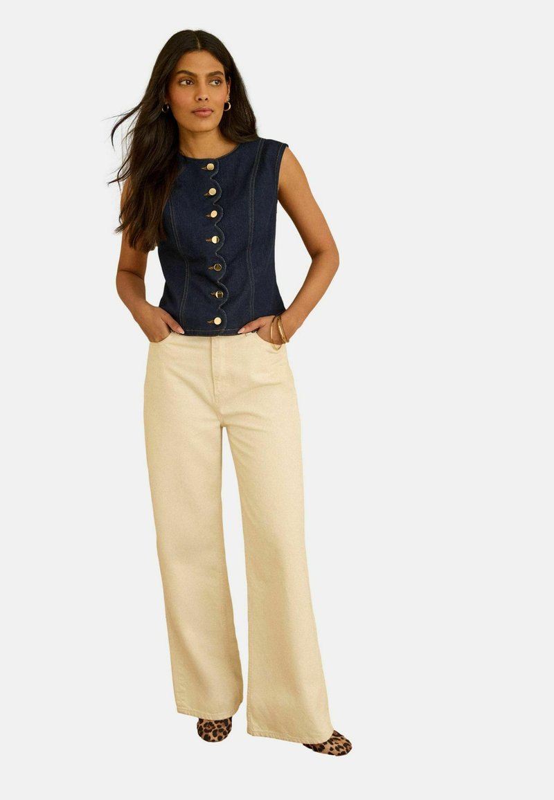 Femeie purtând un top fără mâneci din denim închis la culoare, cu nasturi frontali cu margini ondulate, pantaloni largi bej și pantofi cu imprimeu leopard, stând cu mâinile în buzunare.