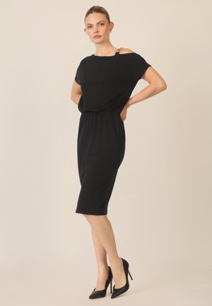Schwarzes Kleid mit lockerer Passform, gerafftem Taillenbereich und kurzen Ärmeln. Auffälliges Detail: goldene Beschläge an der Schulter. Kombiniert mit schwarzen High Heels.
