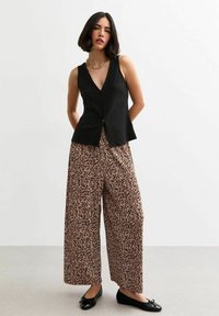 Schwarzes ärmelloses Hemd mit Knöpfen, kombiniert mit weit geschnittenen, hellbraunen Hosen mit einem dunklen Leopardenmuster, ergänzt durch schwarze Ballerinas.