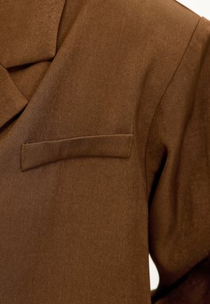 Blazer marron ajusté en tissu texturé ; possède un revers cranté et une poche poitrine simple du côté gauche.