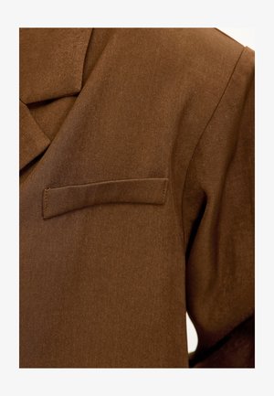 Blazer marron ajusté en tissu texturé ; possède un revers cranté et une poche poitrine simple du côté gauche.