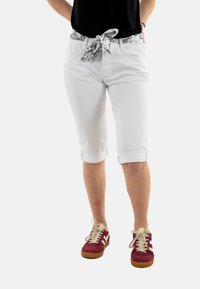 Witte knielange broeken met een zwart-wit gestreepte riem, opgerolde pijpen en voorzakken, gedragen met rood-witte sneakers.