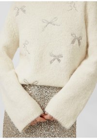 Cremefarbener Pullover mit langen, weiten Ärmeln, verziert mit silbernen Pailletten-Bow-Motiven. Kombiniert mit einem schimmernden, goldenen Paillettenrock. Weiche Textur.