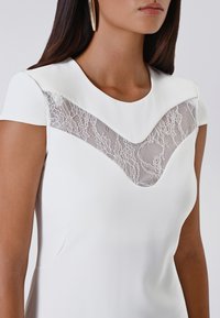 Donna che indossa un vestito bianco con maniche corte e un pannello in pizzo floreale in rete sulla parte superiore del petto e del collo.