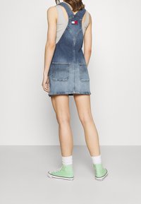 Person som bär en ärmlös grå topp, blå denim overallklänning med bakfickor, vita strumpor och mintgröna högskurningar.