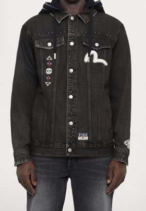 Sort denimjakke med metalknapper, broderede mønstre på venstre bryst og ærme samt en hætte med snore, båret med falmede sorte jeans.