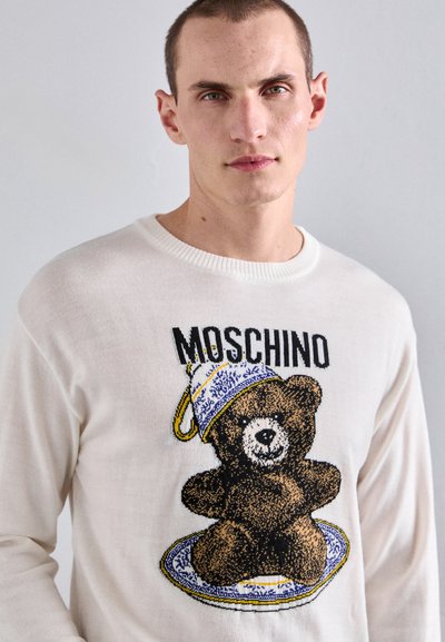 Weißer Strickpullover mit einem Motiv eines braunen Teddybären, der eine blau-weiße Teetasse hält, akzentuiert mit dem Markennamen "Moschino" darüber.