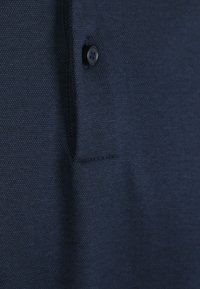 Navyblauwe polo met close-up, met een gestructureerde weefsel, een enkele zwarte knop en een kraag. Het materiaal lijkt ademend en lichtgewicht.