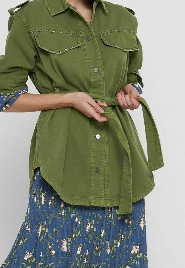 Personne nouant une ceinture sur une veste verte à boutons portée sur une jupe plissée bleue à fleurs, sur fond uni.