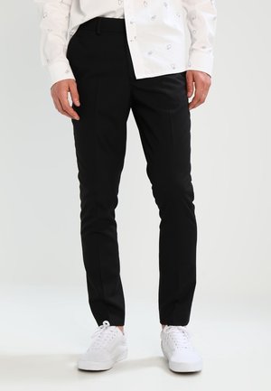 Hombre vistiendo pantalones negros ajustados, zapatillas blancas y una camisa blanca con dibujos de manos en línea negra sobre un fondo claro y sencillo.