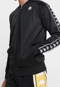 Black zip-up jacket made of smooth fabric, featuring white Kappa logo and dual white stripes with repeated logo along the sleeves.
