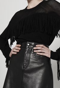 Pull en maille noire avec détails à franges sur l'épaule, associé à une jupe en cuir noire dotée d'une couture frontale et de ferrures métalliques.
