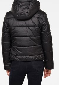 Černá péřová bunda s kapucí, prošívanou texturou a elastickými manžetami, kombinovaná s tmavými džínami. Bunda má lesklý povrch.