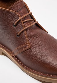 Hnědá kožená bota s texturovaným povrchem, zaobleného tvaru, s dvěma otvory pro šněrování a béžovým prošíváním podél podrážky.