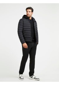 Giacca a vento nera con cappuccio, caratterizzata da cuciture orizzontali, tasche con zip e una silhouette aderente, abbinata a pantaloni neri e sneaker.