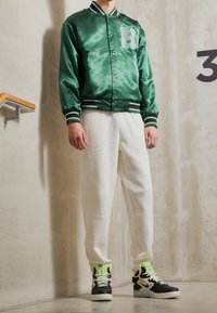 Grüne Satin-Baseballjacke mit weißem "H"-Logo, gestreiften Bündchen, kombiniert mit weißen Hosen und schwarzen Sneakern mit neon-grünen Akzenten.