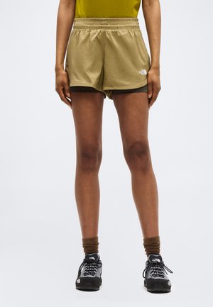Personne portant un short superposé vert olive, des chaussettes marron et des chaussures de randonnée noires et blanches, debout devant un fond clair uni.