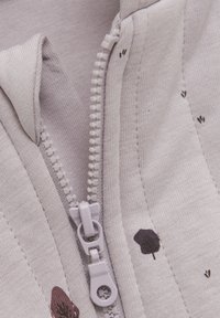Lys grå zip-up hoodie med et struktureret stof, der har en plastisk lynlås og små brune mønstre, der minder om skyer. Nærbillede af lynlåsen.
