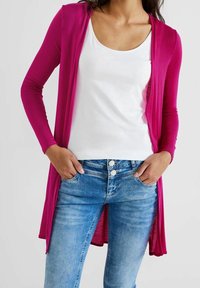 Kvinna i en vit topp med rund halsringning, en ljusrosa lång kofta och ljusblå jeans med händerna i framfickorna.