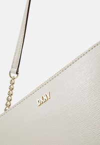 Svetle šedá kabelka DKNY s texturovaným povrchem, zlatým logem a řetízkem. Má zipové uzavření a elegantní úhlový design.