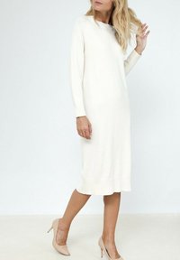 Robe en tricot blanche, longueur genou et à manches longues, avec un ourlet côtelé. Portée avec des chaussures à talons nus, elle présente une coupe décontractée et un décolleté en bateau.