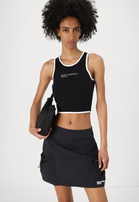 Fekete cropped tank top fehér díszítéssel; logóval ellátott. Párban egy fekete szoknyával, amelynek zsebei és cipzáras részletei vannak. A modell egy fekete táskát tart.