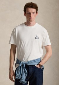 Mladý muž kučeravých vlasov, oblečený v bielom tričku Polo Ralph Lauren, modrých džínsoch a riflej košeli previazané okolo pása.