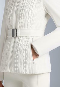 Giacca bianca con design a trecce lavorate a maglia testurizzata, vita aderente e fibbia della cintura argento, con una tasca con cerniera sul lato destro.