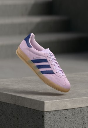 Baskets Adidas Gazelle violet clair avec des bandes bleu marine et une languette au talon, semelle en gomme, flottant au-dessus d’un rebord en pierre avec des escaliers flous en arrière-plan.