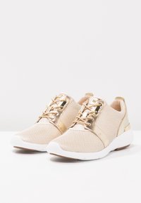 Beżowe siateczkowe trampki z metalicznymi, złotymi akcentami i białymi podeszwami. Sznurowadła są plecione, z gładką teksturą na złotych częściach.