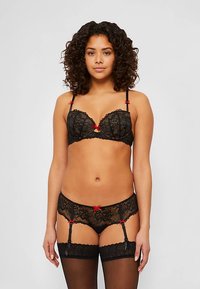 Czarny koronkowy komplet bielizny z ozdobnymi czerwonymi kokardkami, obejmujący biustonosz typu balkonette, dopasowane majtki oraz paski do pończoch. W zestawie półprzezroczyste, czarne pończochy do uda.