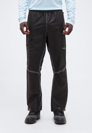 Homme portant un pantalon de pluie noir et des chaussures de randonnée noires, debout devant un fond uni, les mains détendues le long du corps.