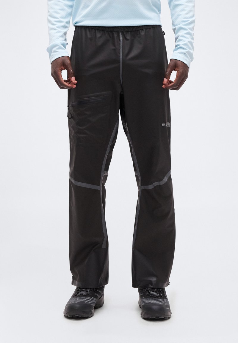 Άνδρας φορώντας μαύρο αδιάβροχο παντελόνι υπαίθρου και μαύρα πεζοπορικά παπούτσια, στέκεται μπροστά σε απλό φόντο, με τα χέρια χαλαρά στα πλευρά.