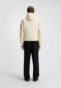 Giubbotto imbottito con cappuccio, di un beige chiaro, con una silhouette corta, indossato sopra pantaloni neri a gamba larga. Texture liscia, dettagli minimi.