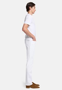 Białe dżinsy o slim fit, noszone z dopasowaną białą koszulą i brązowymi zamszowymi botkami za kostkę, przedstawione w widoku bocznym.