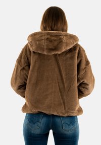 Chaqueta con capucha de pelo sintético marrón, con una textura suave, un corte oversized y mangas largas, usada sobre unos jeans de mezclilla azules con un diseño de bolsillo distintivo.