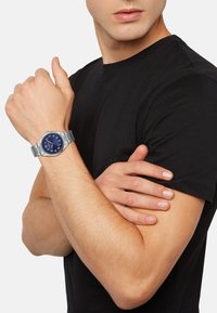 Montre en métal argenté avec un cadran bleu, de grands chiffres et un bracelet à maillons, portée sur un avant-bras contre un fond de chemise noire.