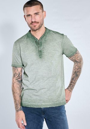 Mann mit kurzen Haaren und Tätowierungen an beiden Armen trägt ein grünes Henley-Shirt mit kurzen Ärmeln und blaue Jeans, posiert mit einer Hand auf der Hüfte.
