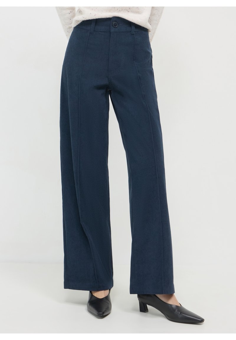 Personne portant un pantalon large en velours côtelé bleu marine et des chaussures noires à talons bas et bouts pointus, debout devant un fond uni.