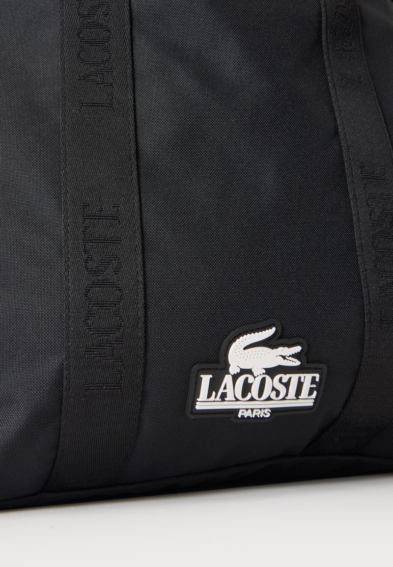 Borsa in tessuto nera con logo Lacoste in rilievo sulle cinghie e patch con logo che mostra un coccodrillo bianco e il testo Lacoste Paris.