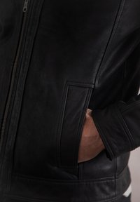 Black leather jacket with a fitted design, featuring a smooth texture and a side pocket. Zipper closure and tailored cuffs are visible.