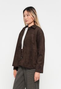 Ung kvinne med langt blondt hår iført en brun knappet jakke, hvit topp og grå vide bukser, stående sideveis mot en hvit bakgrunn.