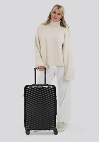 Frau in beige Rollkragenpullover und weißen Hosen steht und hält den Griff eines schwarzen Hartschalenreisekoffers mit vier Rädern.