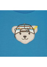 Tela azul con el rostro de un oso bordado que lleva una gorra. La gorra tiene rayas horizontales en naranja, amarillo y azul. Detalle texturizado en las características del oso.