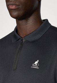 Svart polo-skjorte med glidelås, med en smal krage. Inkluderer detaljer med hvit logo på venstre brystområde. Glatt tekstur.