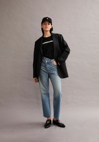 Blazer negro sobre una camiseta gráfica negra, jeans de cintura alta en azul claro y zapatos negros sin cordones, con una gorra negra que presenta un logotipo.