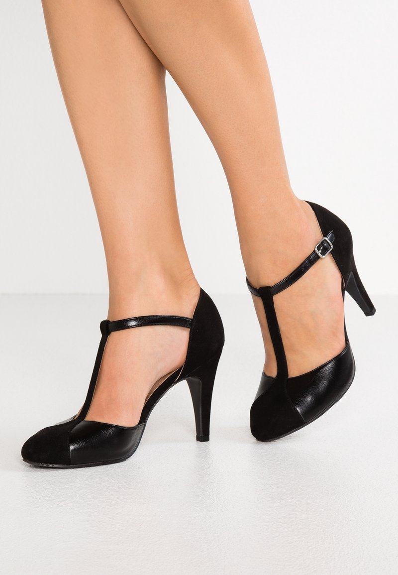 Jambes portant des escarpins noirs à bride en T avec un talon stiletto de hauteur moyenne sur un fond de sol blanc.