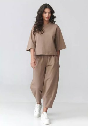 Kobieta o długich kręconych włosach, ubrana w luźną beżową bluzkę i spodnie oraz białe trampki, spacerująca na tle jednolitego jasnego tła.